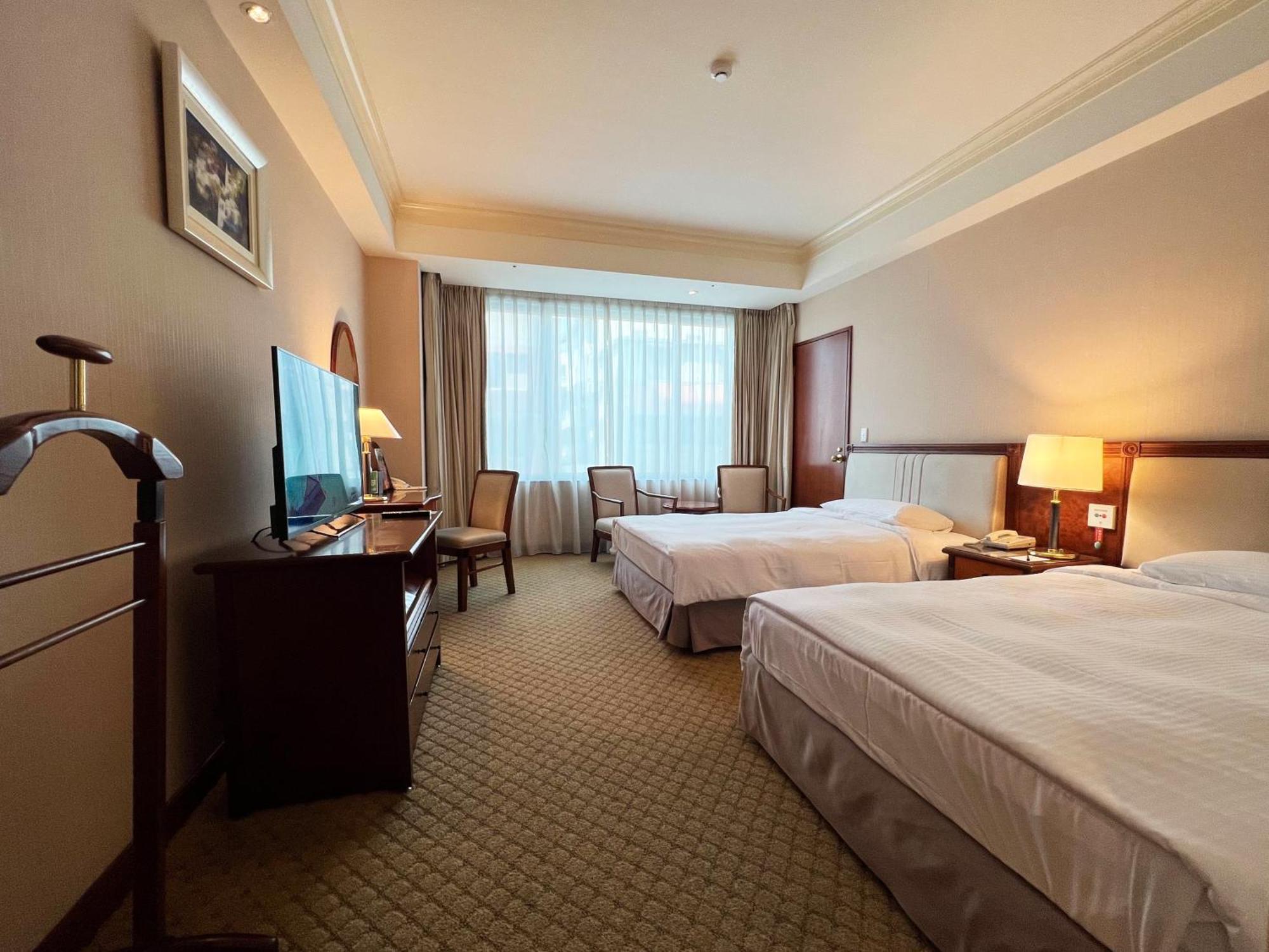 Evergreen Laurel Hotel - Taichung Exterior foto The photo shows a hotel room featuring two twin beds placed parallel to each other. The room has a warm color palette and includes a large window with sheer curtains allowing natural light to illuminate the space. There is a wooden desk and two chair